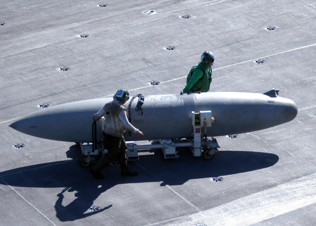 Zwei Männer in Schutzausrüstung inspizieren eine große zylindrische Rakete mit spitzer Nase auf einem Flugzeugträger.