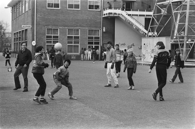 Eine Gruppe von Menschen, die vor einem Gebäude mit Fenstern, Treppen mit Geländern und einem Laternenmast Fußball spielen, mit Bäumen im Hintergrund, in Schwarz-Weiß.