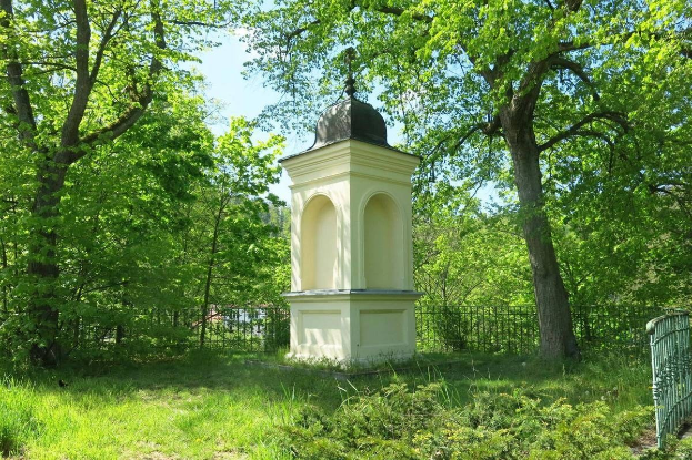 Kleines weißes Holocaust-Gedenkmonument in einem grasbewachsenen Friedhof umgeben von einem Zaun und Bäumen unter einem klaren blauen Himmel in Vilnius, Litauen.