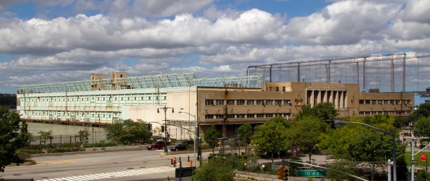 Eine breite Stra├če mit Fahrzeugen, ges├Ąumt von B├Ąumen und Stra├čenlaternen, f├╝hrt zu einem gro├čen Geb├Ąude in der N├Ąhe eines Gew├Ąssers.