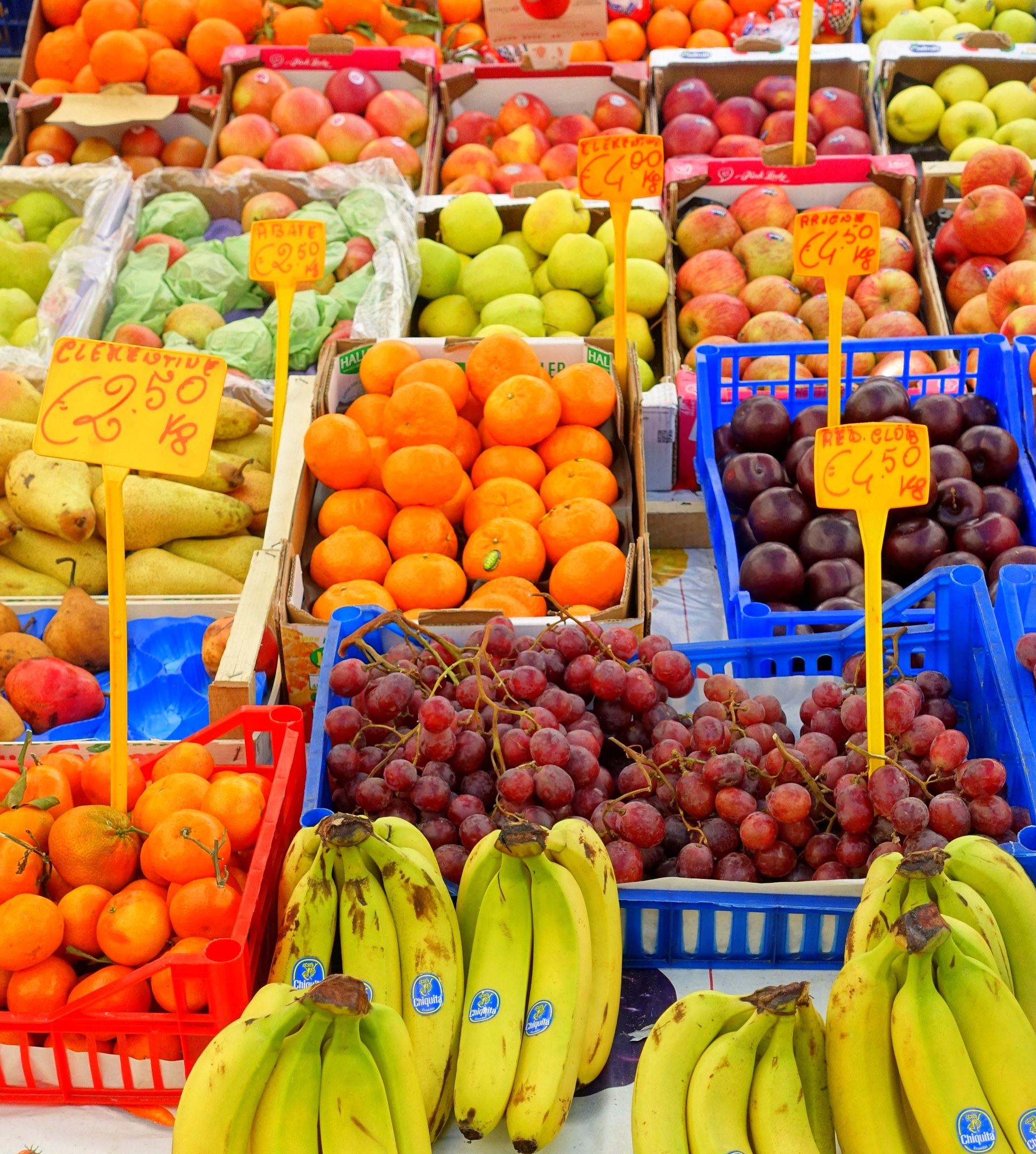 Ein Marktstand mit verschiedenen Obst- und Gemüsesorten und daran befestigten Preisschildern.