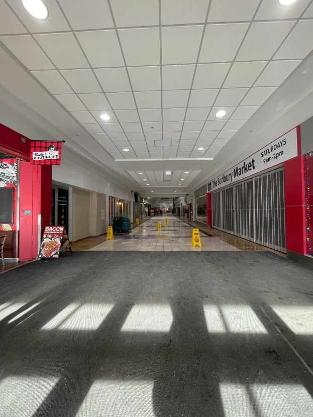 Innenansicht eines hell erleuchteten Einkaufszentrums mit einem Stuhl und einer Tafel, die "Sydbury Market - Vorher und Nachher" links zeigt, und verstreuten Gegenständen rechts und auf dem Boden.