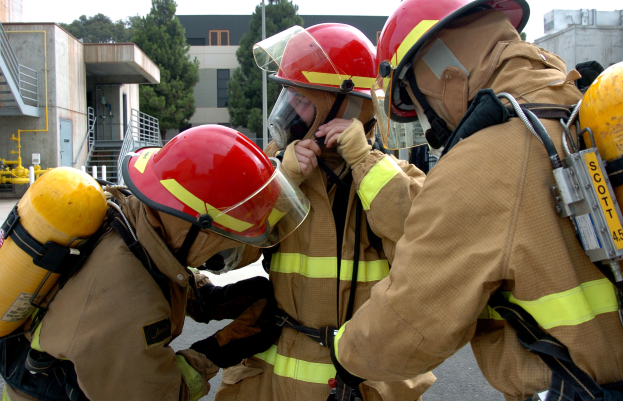 Drei Feuerwehrleute in voller Ausrüstung, einschließlich Helmen und Sauerstoffflaschen, stehen gemeinsam vor einem Gebäude, mit anderen Gebäuden, Bäumen und Polen im Hintergrund unter einem klaren blauen Himmel.
