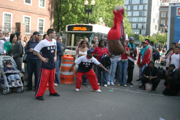 Eine Menschenmenge, einige mit Mützen, ein Kind im Kinderwagen, ein Luftballon und ein Verkehrsobjekt im Vordergrund, ein Bus, Bäume, Gebäude mit Fenstern, ein Laternenpfahl und ein Geländer im Hintergrund.