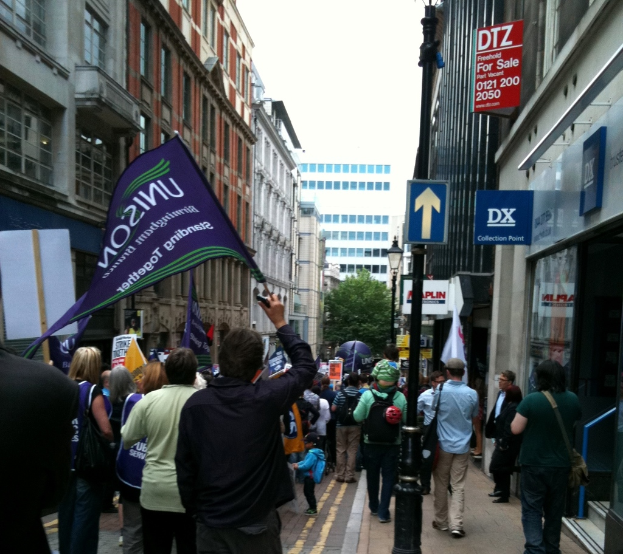 Eine große gruppe von menschen auf einer straße, einige haltend flaggen und plakate, mit einer schildtafel auf einem pfahl, gebäude mit fenstern an den seiten, und einem baum, leuchtpfahl, gebäude und himmel im hintergrund.