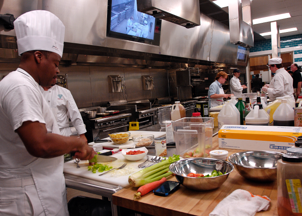 Eine Gruppe von Köchen in einer Küche bei der Zubereitung von Speisen auf einem Schneidbrett, wobei ein Koch im Vordergrund in einer weißen Schürze und einer weißen Mütze steht und ein Messer hält und von verschiedenen Utensilien, Schüsseln, Gemüse, einem Handy, Flaschen und anderen Gegenständen umgeben ist, mit einem Fernseher und Deckenlampen im Hintergrund.