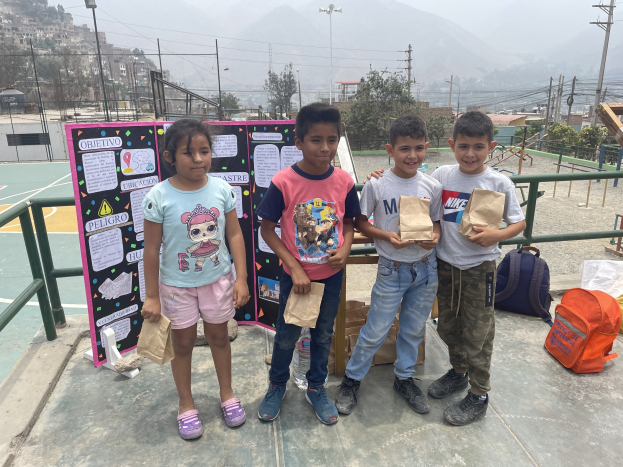 Gruppe von Kindern auf einem Basketballfeld mit Papiertüten in den H├Ąnden, mit Tüten und Gegenst├Ąnden rechts und einer Tafel mit Text links, vor einem Hintergrund aus Zaun, Masten, Laternen, Strommasten, B├Ąumen, Geb├Ąuden, H├Ąngen und Himmel.