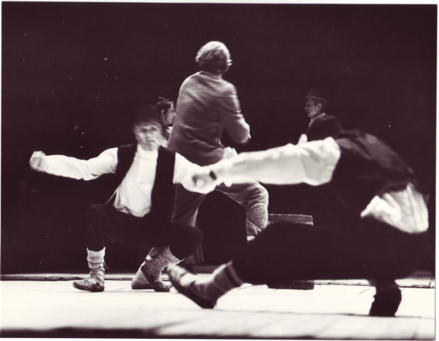 Eine Gruppe von Menschen, die auf einer Bühne tanzen, in einem Schwarz-Weiß-Foto, mit einer Person in der Mitte, die Schuhe trägt, vor einem dunklen Hintergrund.