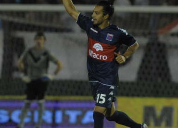 Ein Fußballspieler feiert nach einem Tor auf dem Boden, mit einer jubelnden Menge und einer Person im Hintergrund, alles mit einem unscharfen, energetischen Hintergrund.