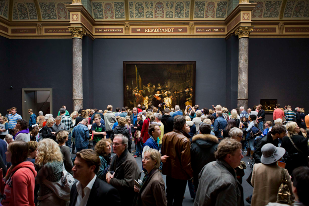 Eine große Gruppe von Menschen steht in einem Raum mit einem Foto Rahmen an der Wand, Säulen, einer Tür und einer Decke mit Lampen, einige halten Taschen, im Museum of Fine Arts in Amsterdam.