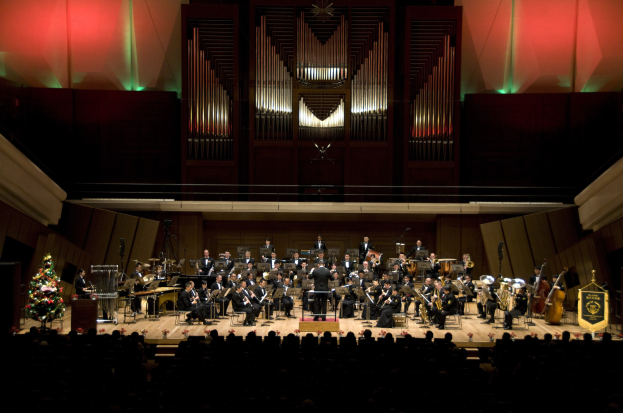 Großes Orchester auf der Bühne mit einem Weihnachtsbaum im Hintergrund, Musiker sitzend mit Instrumenten, Dirigent am Podium und Texttafeln auf der rechten Seite.