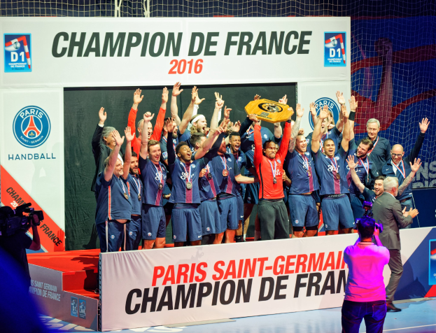 Eine Gruppe von Männern auf einer Bühne mit einem Pokal, einer Person, die filmt, und einem "Champion de France 2016"-Schild im Hintergrund.