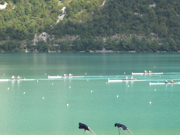 Gruppe von Menschen beim Ruderwettbewerb auf einem Gewässer mit Bäumen und einem Hügel im Hintergrund, Kleidung auf Ständen darunter.