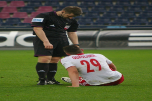 Ein Fußballspieler sitzt neben einem Schiedsrichter auf dem Boden in einem Stadion, beide tragen Sportkleidung, mit Schildern und Stühlen im Hintergrund.