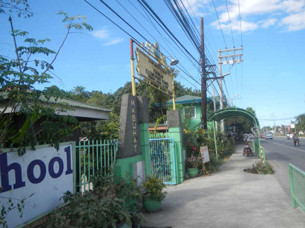 Der Eingang einer Schule mit einem Schild, das "Schule" trägt, an der Seite einer Straße, mit einem Bogen, Tor, Namensschild, Pflanzen, Bäumen, Strommasten mit Drähten und einem bewölkten Himmel im Hintergrund.