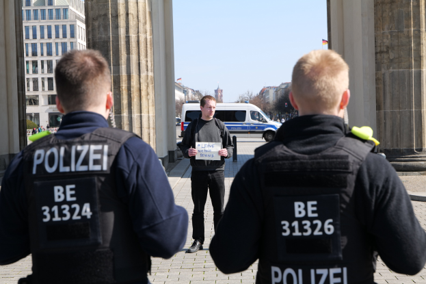 Drei Polizeibeamte stehen vor einem Gebäude, mit einem Mann, der ein Papier zwischen ihnen hält, flankiert von Säulen, während Fahrzeuge, Menschen, Bäume und Gebäude den Hintergrund füllen.