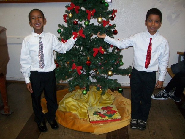 Ein geschmückter Weihnachtsbaum steht in einem Raum mit einer Kiste und anderen Gegenständen auf dem Boden, einem Holztisch links, einem Stuhl rechts und zwei Jungen in weißen Hemden, die strahlend den Baum halten.