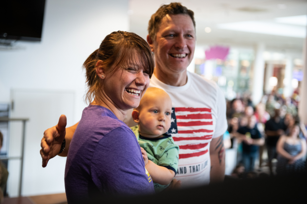 Ein Mann in einem weißen T-Shirt und eine Frau mit einem Baby stehen vor einer Menge, mit einem Fernseher an der Wand im Hintergrund.