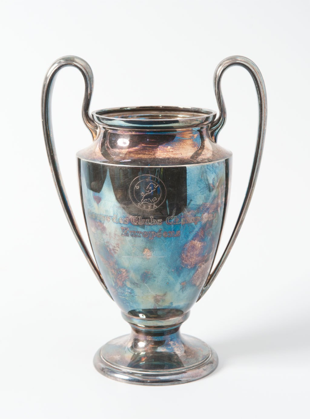 Der UEFA-Champions-League-Pokal, ein stählerner Becher mit zwei Griffen und einem blauen und silbernen Farbschema, beschriftet mit "UEFA Champions League", vor einem weißen Hintergrund.