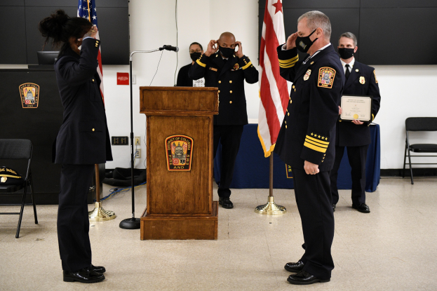 Gruppe uniformierter Personen in Masken vor einem Rednerpult mit Mikrofon, umgeben von Flaggen und Möbeln, bei einer Vereidigung eines neuen Feuerwehrchefs