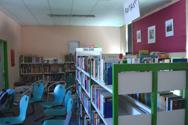 Eine Bibliothek mit Bücherregalen voller Bücher, Stühlen, Taschen auf dem Boden, gerahmten Bildern an den Wänden, einer Tafel mit Text, einer Tür und Deckenleuchten.