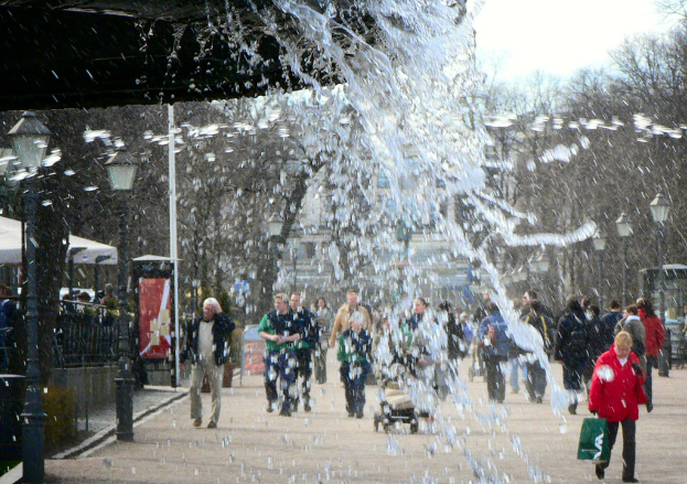 Wasser läuft aus einer unsichtbaren Quelle oben, mit einer Gruppe von Menschen, die unten auf der Straße gehen, Bäumen auf der rechten Seite und Straßenlaternen auf der linken Seite.