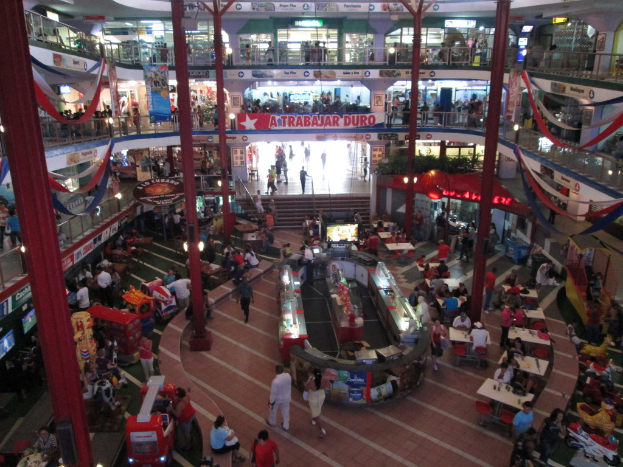 Ein belebtes Einkaufszentrum-Innenbild mit Säulen, Menschen, Tischen, Fliesenboden, Flaggen, Geschäften, einem Banner, Treppen und einem Namensschild.