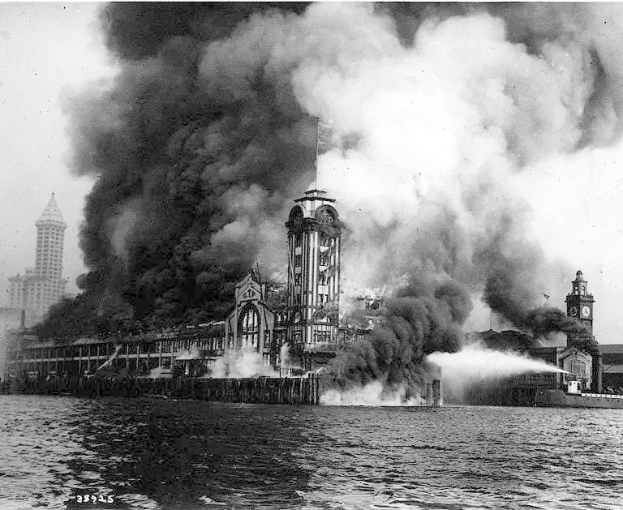 Schwarz-weiß-Foto eines großen, von Flammen eingehüllten Gebäudes mit Rauchwolken, einem Gewässer im Vordergrund und anderen Gebäuden und Himmel im Hintergrund.