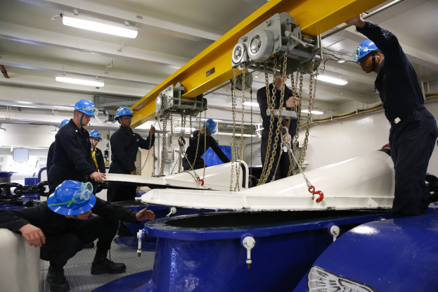 Eine Gruppe von Männern in Helmen arbeitet an einem Boot, umgeben von Maschinen und Equipment, mit einer Wand und Deckenbeleuchtung im Hintergrund, was darauf hindeutet, dass sie eine Windkraftanlage installieren.