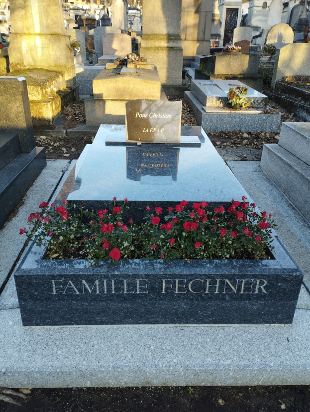 Ein Friedhof mit einem zentralen Grab, geschmückt mit roten Blumen, umgeben von Bäumen unter einem klaren blauen Himmel.