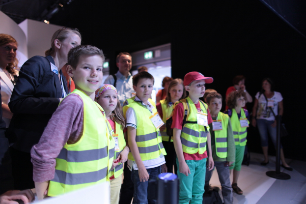 Eine Gruppe von Kindern mit Sicherheitswesten und Namensschildern, mit einem Jungen, der links lächelt, auf einem Boden mit einem Pfahl rechts und einigen Menschen im Hintergrund.