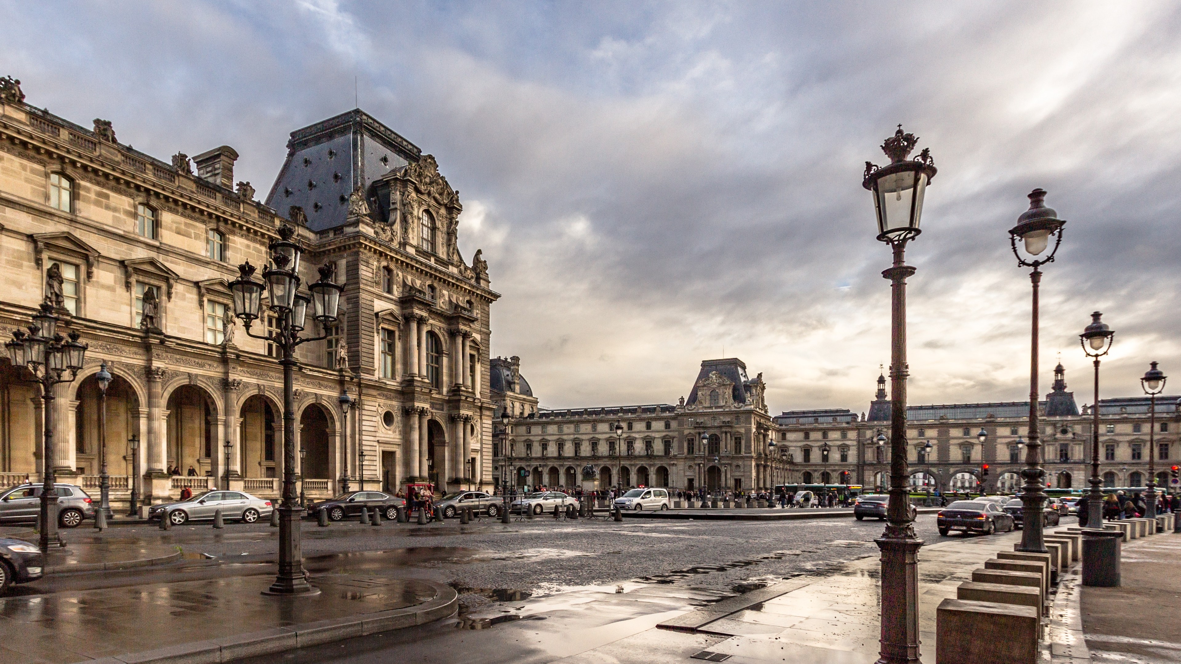 Das Louvre-Museum in Paris, Frankreich, mit seinen ikonischen Gebäuden, Straßenlaternen, Straßenschildern, Kraftfahrzeugen auf der Straße, Fußgängern auf dem Gehweg und einem bewölkten Himmel.