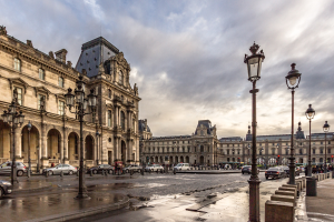 Das Louvre-Museum in Paris, Frankreich, mit seinen ikonischen Gebäuden, Straßenlaternen, Straßenschildern, Kraftfahrzeugen auf der Straße, Fußgängern auf dem Gehweg und einem bewölkten Himmel.