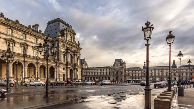 Das Louvre-Museum in Paris, Frankreich, mit seinen ikonischen Gebäuden, Straßenlaternen, Straßenschildern, Kraftfahrzeugen auf der Straße, Fußgängern auf dem Gehweg und einem bewölkten Himmel.