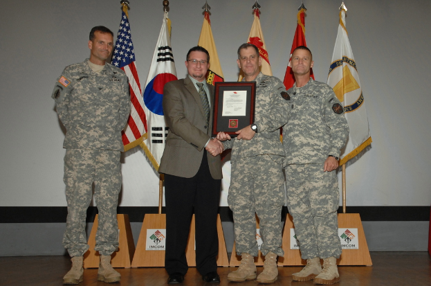 Vier Männer stehen auf einer Bühne: drei in Armeeuniformen und einer in Blazer, Krawatte und Hemd, die Hände schütteln und einen Preis überreichen, mit sechs Fahnen auf Ständen und einer weißen Wand im Hintergrund.