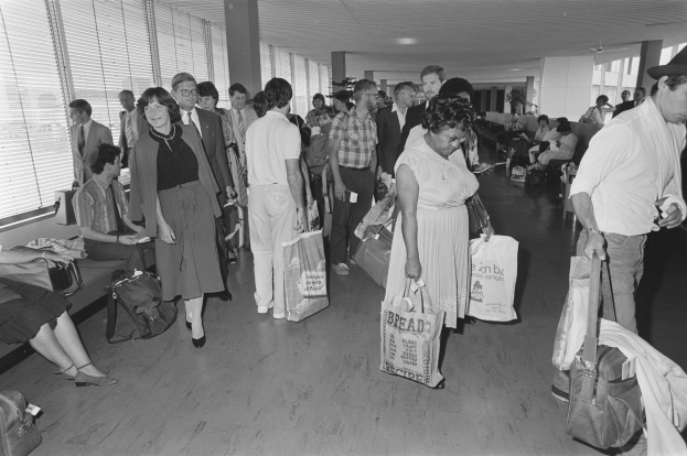 Ein Schwarz-Weiß-Bild von Reisenden in einem Flughafen, einige sitzen auf Bänken und andere stehen, mit einem Mann und einer Frau, die Taschen halten, in der Mitte und Jalousien links.