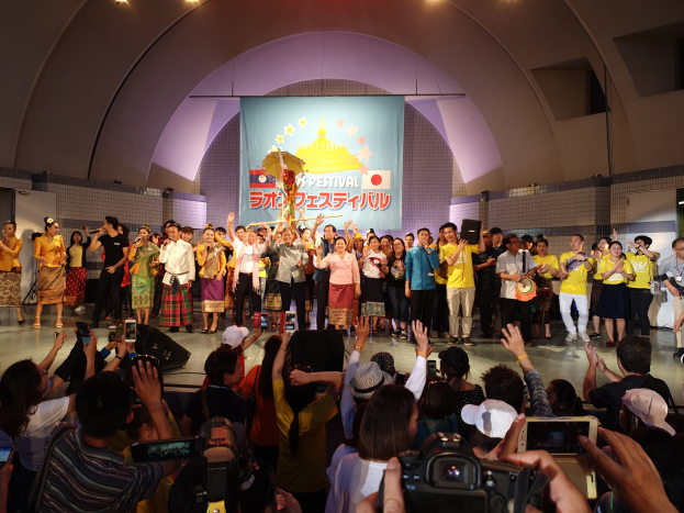 Gruppe von Menschen auf einer Bühne mit Kameras, Handys und Musikinstrumenten während einer Festivaleröffnung, mit einem Banner und Deckenbeleuchtung im Hintergrund.