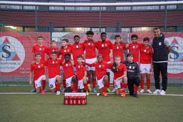Eine Gruppe junger Männer in roten T-Shirts und schwarzen Shorts steht auf einem Fußballfeld vor einer Kiste roter Gegenstände, im Hintergrund sind Banner mit der Aufschrift "FC Red Black Eglaute - Champions League" zu sehen.