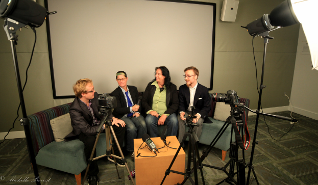 Eine Gruppe von drei Personen sitzt auf einem Sofa und gibt ein Interview, wobei eine Person links in einem schwarzen Mantel und Hose, eine Person in der Mitte mit einem Mantel und blauer Krawatte und einem Mikrofon sowie eine Person rechts mit einem Lächeln zu sehen ist. Ein Mikrofonrecorder befindet sich auf dem Tisch in der Mitte, Kameras sind auf der rechten und linken Seite und große Scheinwerfer sind vorhanden, sowie ein Projektionsschirm im Hintergrund.