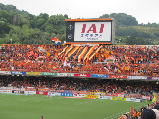 Ein Fußballspiel wird in einem Stadion mit einer großen Zuschauermenge, grünem Rasen, einem Tor, Bannern, Fahnen, einem großen Bildschirm, Bäumen und einem klaren blauen Himmel gespielt.