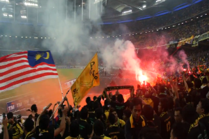 Eine große Menschenmenge in einem Stadion mit Bengalos, Fahnen und einer Kamera, mit Rauch und Schildern, die "Malaysia vs Malaysia Live Streaming" im Hintergrund anzeigen.