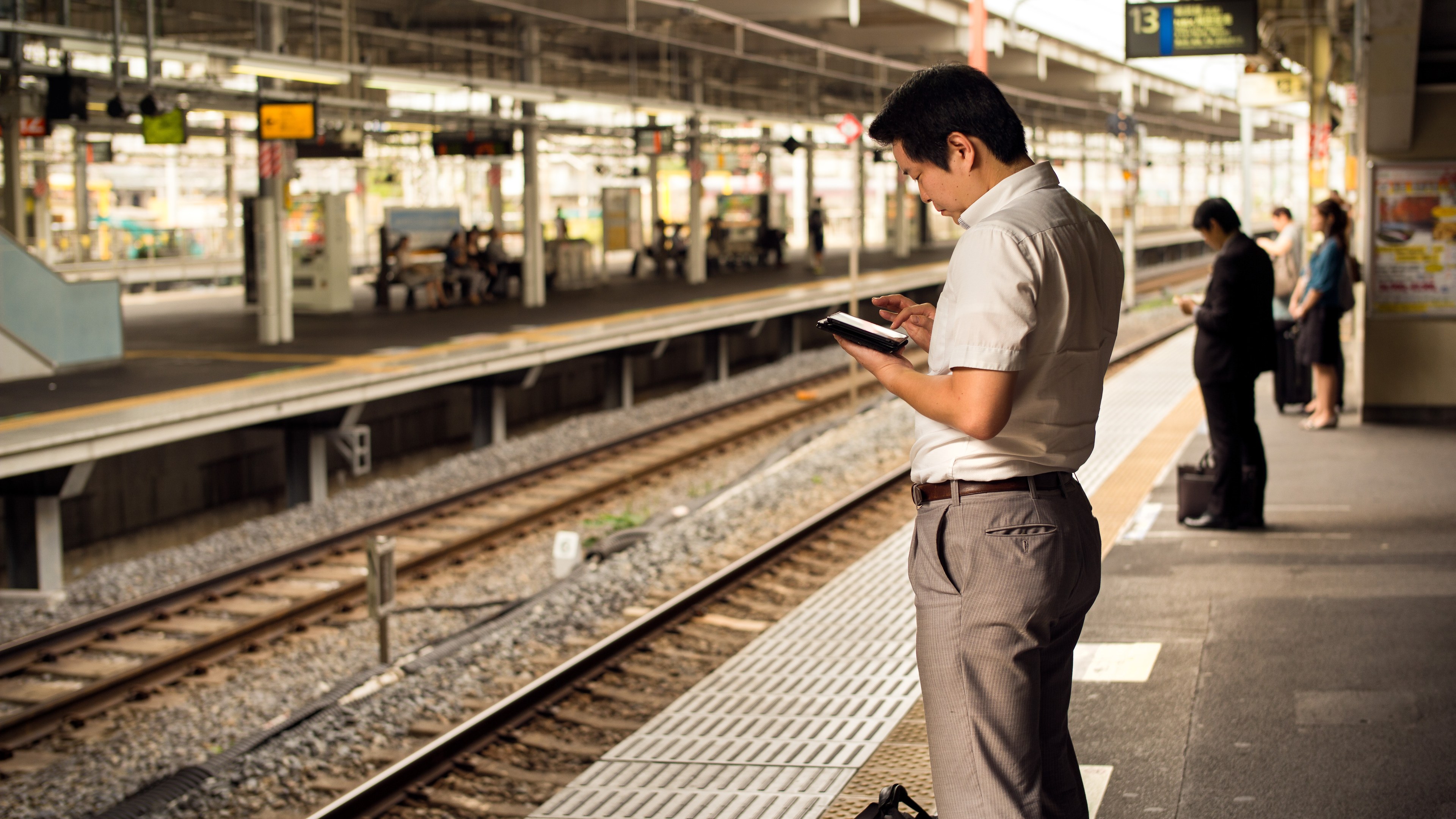Ein Mann steht auf einem Bahnsteig, sieht auf sein Handy, umgeben von anderen Menschen mit Bahnschienen im Hintergrund; ein Schild mit Text ist auf der rechten Seite zu sehen.
