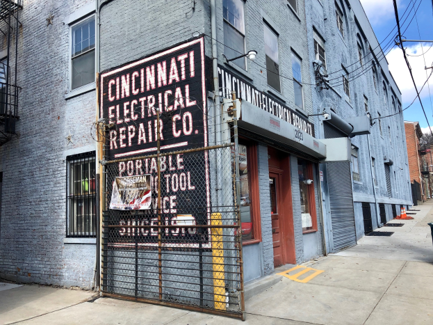 Außenseite des Gebäudes der Cincinnati Electrical Repair Co. mit einem Schild, einem Metallzaun, einem Müllcontainer, Strommästen, Drähten, Bäumen und einem bewölktem Himmel.