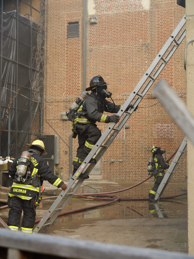 Feuerwehrleute in Helmen und Ausrüstung klettern an einer Leiter vor einem Backsteingebäude mit Rohren auf dem Boden und einer Metallstange unten, mit einem anderen Gebäude und einem Netz im Hintergrund.