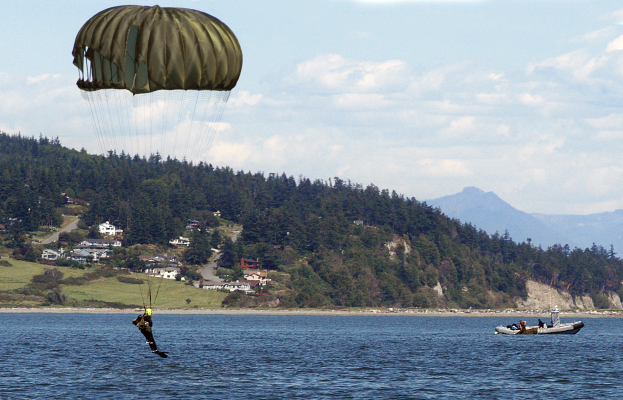 Eine Person gleitschirmfliegt über einen See mit einem Boot auf der rechten Seite, Bäume, Gebäude, Hügel und einen klaren blauen Himmel im Hintergrund.