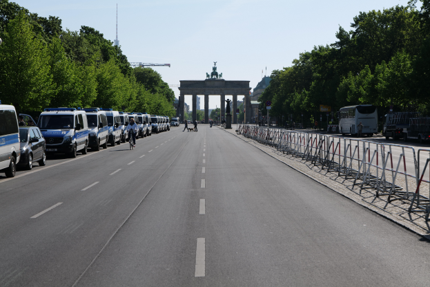 Eine Reihe von Polizeiwagen auf einer Straße vor dem Brandenburger Tor in Berlin, Deutschland, mit Menschen auf Fahrrädern und in der Nähe Stehenden, Barrieren, Bäumen und einem Bogen mit Statuen im Hintergrund.