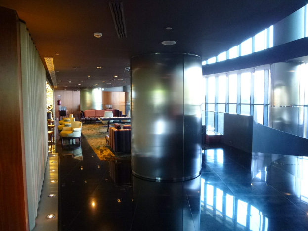 Eine geräumige First-Class-Lounge am Hong Kong International Airport mit großen Fenstern, hohen Decken mit elektrischem Licht, Sitzanordnungen einschließlich Stühlen und Sofas, Tischen, einem Teppichboden und einer mittigen Säule.