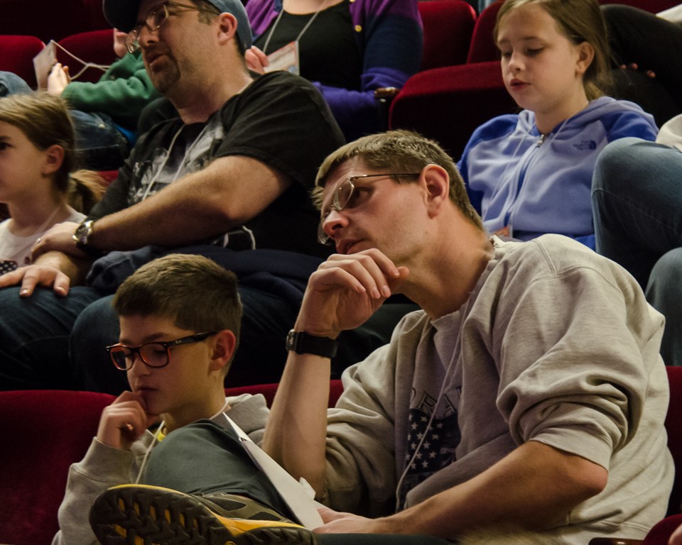 Eine Gruppe von Menschen sitzt in einem Kino, mit einem Mann und einem Jungen mit Brille in der Mitte.