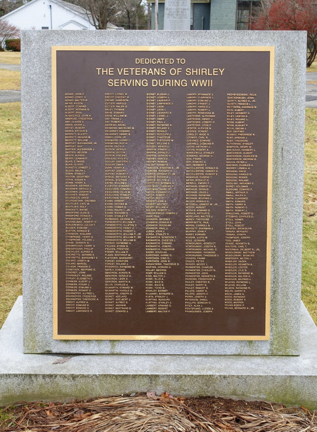 Ein Gedenkstein mit einer Tafel, auf der "Veteranen von Shirley im Zweiten Weltkrieg" steht, befindet sich vor einem Gebäude, umgeben von Gras und trockenen Blättern, mit Bäumen und einem klaren blauen Himmel im Hintergrund.