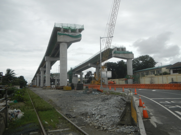 Baustelle mit einer Brücke im Hintergrund, Straße mit Absperrkegeln, verstreute Steine und Gras, Eisenbahnschiene links, Bäume und Gebäude auf beiden Seiten und bewölkter Himmel.